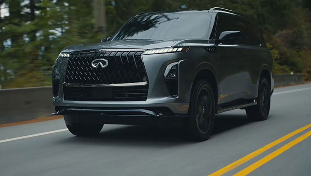 A dark gray Infiniti QX80 SUV with a large black grille driving on a winding two-lane road lined with trees.